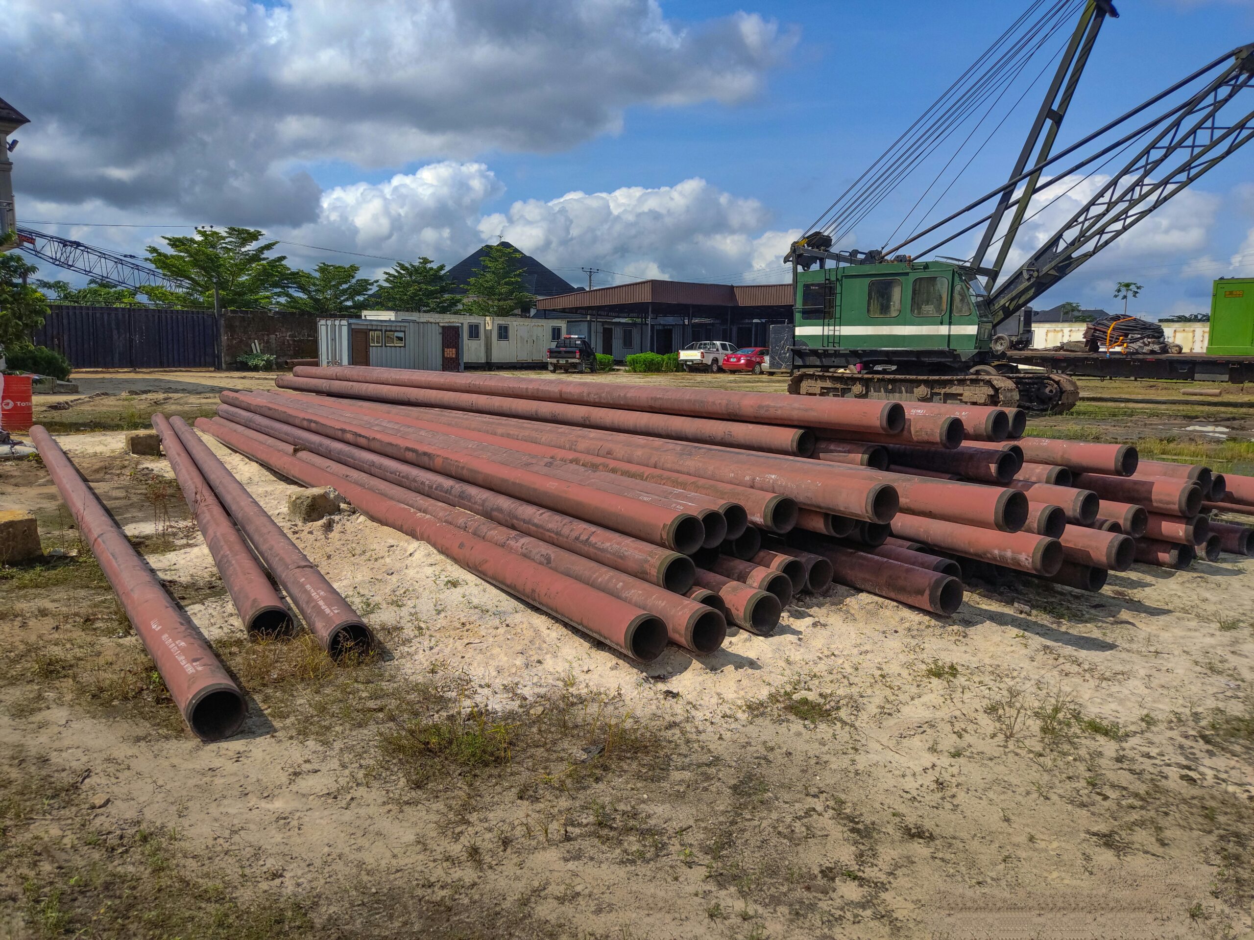 A construction site in Warri, Nigeria featuring stacked rusty metal pipes and heavy machinery.